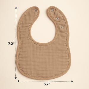May include: A tan baby bib, measuring 7.2 inches long and 5.7 inches wide, with a curved bottom and snap closures at the neck. The bib has a stitched border and is made of a soft, textured fabric.