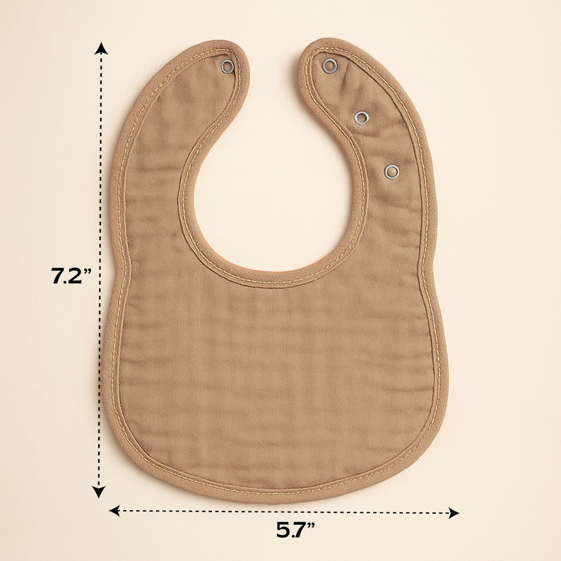 May include: A tan baby bib, measuring 7.2 inches long and 5.7 inches wide, with a curved bottom and snap closures at the neck. The bib has a stitched border and is made of a soft, textured fabric.