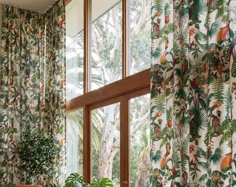 Puede incluir: Cortinas blancas con un estampado de jungla vibrante que presenta monos, loros y follaje exuberante. Las cortinas enmarcan una gran ventana con vista a árboles y un interior luminoso con una silla de cuero y plantas.