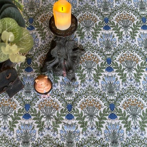 May include: A white tablecloth with a repeating floral pattern in shades of blue, green, and yellow. A lit candle in a black candlestick and a small ceramic pig figurine are on the tablecloth.