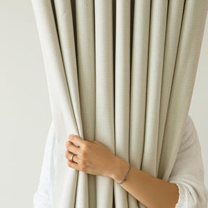 May include: A person's hand reaches out from behind a set of white curtains, holding a silver bracelet.