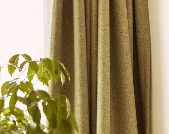May include: Olive green textured curtain panels hanging near a window. The fabric has a subtle woven pattern. A leafy green plant is in the foreground, adding a natural element to the scene. The curtains are gathered, creating soft folds.