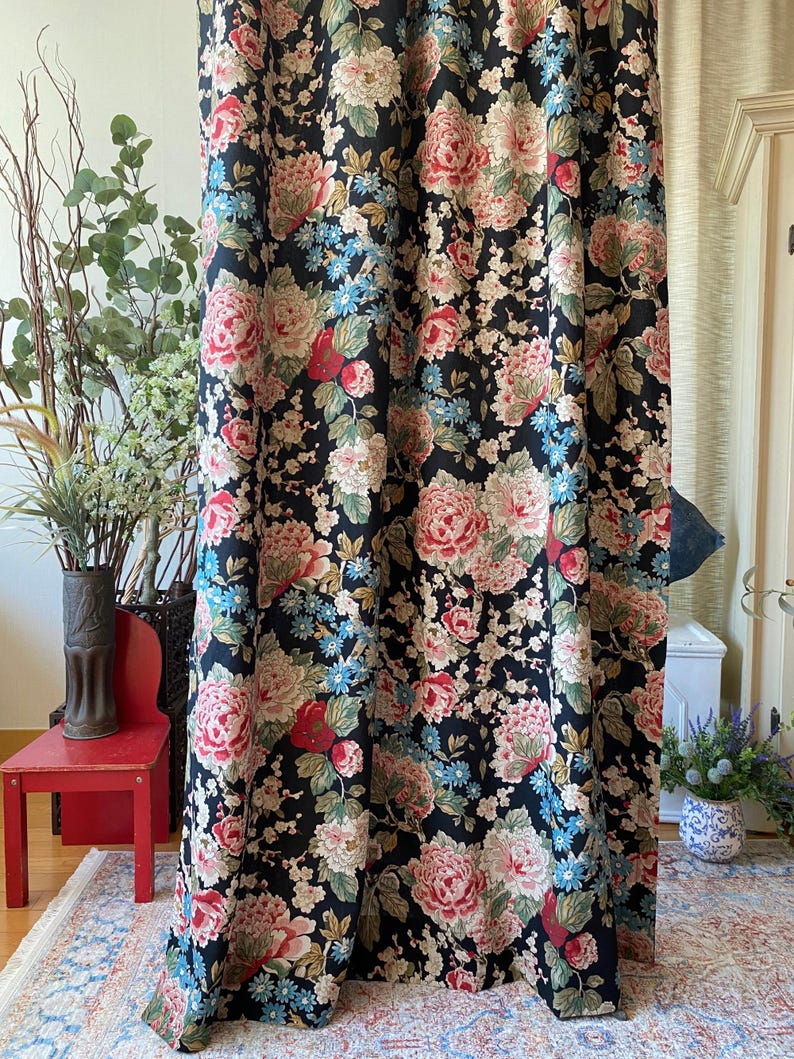 May include: Black floral curtains with large pink, red, and cream-colored peony blooms and smaller blue flowers. The curtain fabric has a dark background and is hanging in front of a window. The curtains are a decorative home decor item.