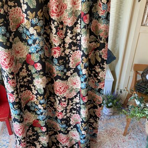 May include: Black floral curtain with large pink, red, and white peony-like flowers, blue accents, and green leaves. The curtain is made of a dark fabric and is hanging in front of a window. The floral pattern is repeated throughout the curtain.