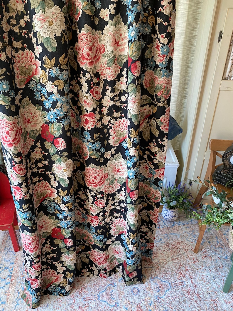 May include: Black floral curtain with large pink, red, and white peony-like flowers, blue accents, and green leaves. The curtain is made of a dark fabric and is hanging in front of a window. The floral pattern is repeated throughout the curtain.
