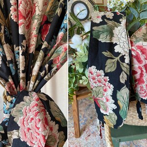 May include: Black floral fabric with large pink, red, and cream-colored peony blooms and green leaves. The fabric is tied in a knot, and also appears as a decorative tie. The fabric is likely used for curtains or home decor.