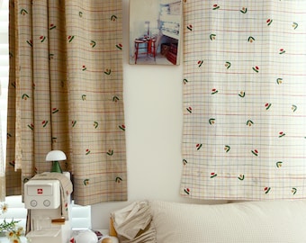 May include: A bedroom with a white wall, a window with beige curtains with a red and green floral pattern, a wooden table with a white coffee maker, a white lamp, and a wooden chair with a beige pillow and a clock on the bed.