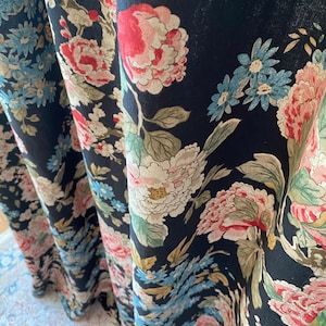 May include: Black fabric with a floral pattern featuring large pink, red, and white peonies, along with smaller blue and white flowers. Green leaves and stems are interspersed throughout the design. The fabric is draped, showing the pattern's repeat.