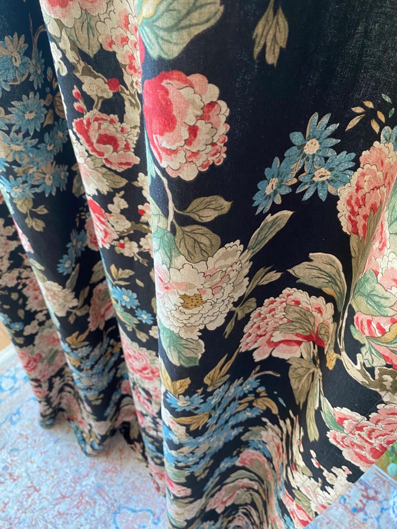 May include: Black fabric with a floral pattern featuring large pink, red, and white peonies, along with smaller blue and white flowers. Green leaves and stems are interspersed throughout the design. The fabric is draped, showing the pattern's repeat.