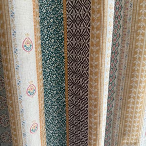 Könnte beinhalten: Nahaufnahme eines gemusterten Stoffes mit vertikalen Streifen. Das Design umfasst Streifen in Weiß mit Blumen- und Paisley-Motiven, Dunkelgrün mit kleinen weißen Blumen, braune Rautenmuster und goldene Streifen mit floralen Details. Der Stoff hat eine Vintage-Ästhetik.
