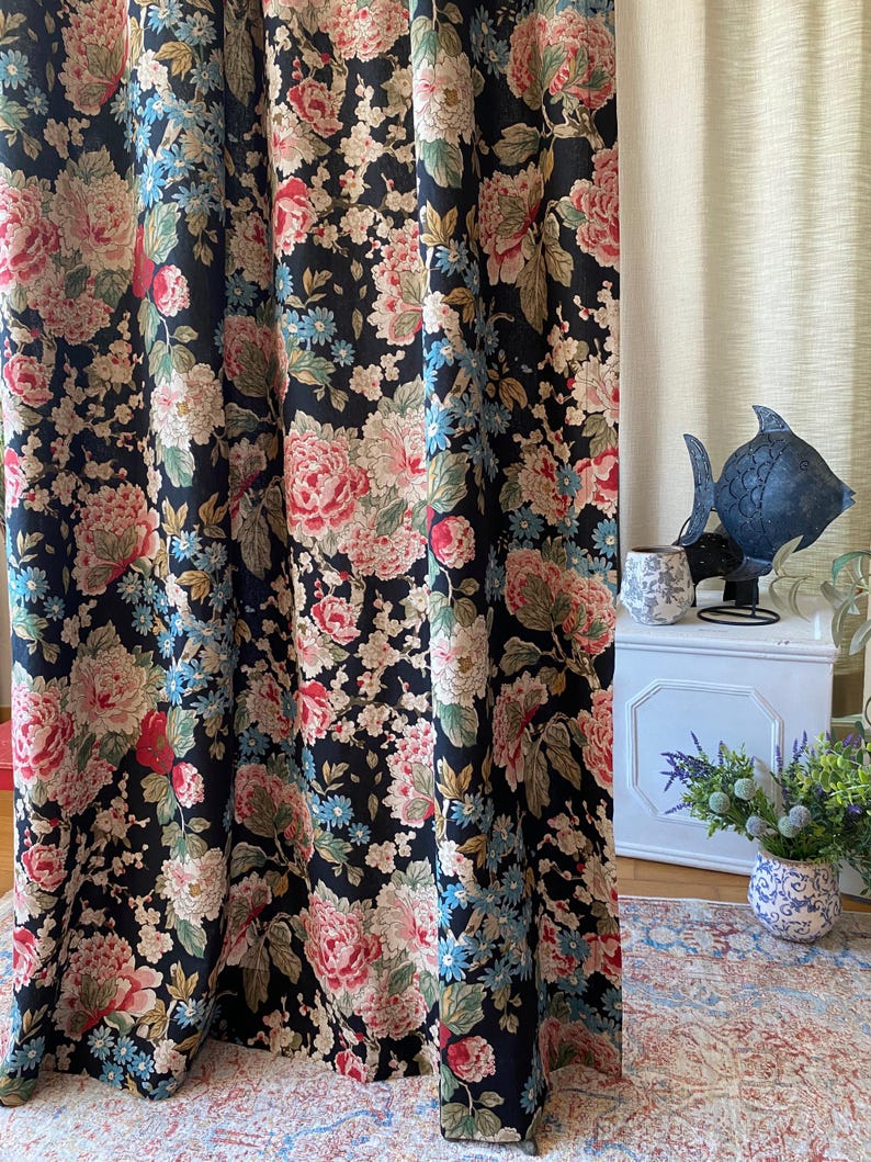 May include: Dark navy floral curtains with large pink and cream peony blooms, accented by small blue flowers and green leaves. The curtains are hanging in front of a neutral-colored wall. The floral pattern is repeated throughout the fabric.