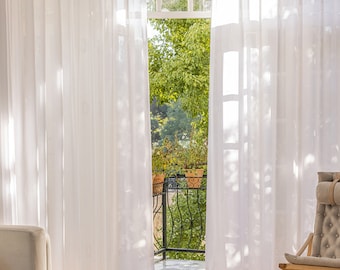 May include: White sheer curtains hang in a living room with a view of a balcony and trees. A white couch, a coffee table with a book and a basket, and a chair are in the room.