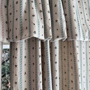 May include: Beige and white striped curtains with a repeating pattern of small red and green floral clusters. The curtains have a vertical stripe design with a light blue border. The fabric is gathered at the top, creating soft folds.