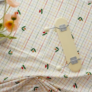 以下が含まれることがあります： 赤、黄、青、緑の格子模様の白いテーブルクロス。テーブルクロスは、小さくカラフルな花の模様で飾られています。クリーム色の木製ハンガーに白いノブが2つ付いており、テーブルクロスの上に置かれています。