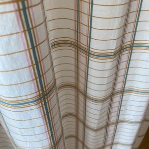 以下が含まれることがあります： 格子柄の白い生地のクローズアップ。格子柄のデザインは、金、ティール、ピンクの色合いの細い線で構成され、格子状のパターンを作り出しています。生地はドレープされており、柔らかい折り目と質感が示されています。