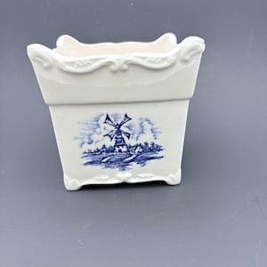 May include: A white ceramic planter with a blue and white windmill scene. The rectangular planter has decorative scrollwork around the top edge and at the corners. The blue windmill scene is on the front of the planter.