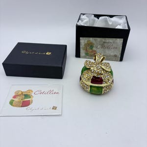 May include: A decorative trinket box in the shape of a gift, with gold-colored accents and sparkling rhinestones. The box is green and red, with a gold bow on top. The box is next to a black box and a card that says "Cotillion".