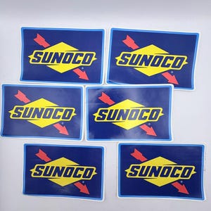 May include: Six rectangular stickers displaying the "SUNOCO" logo. The logo is set against a dark blue background with a yellow diamond shape in the center. Red arrows and white borders complete the design.