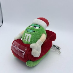 May include: A green plush M&M's character wearing a Santa hat and white gloves, resting in a red plush sleigh. The sleigh has the text "PullString" on the front. A white tag with "m&m" is attached.