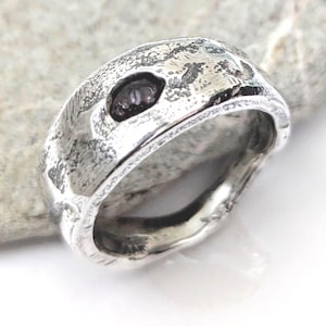 Puede incluir: Un anillo de plata texturizado con una piedra oscura de forma ovalada engastada en la banda. El anillo tiene un aspecto martillado, con una superficie rugosa e irregular. El anillo se muestra sobre una superficie gris claro.