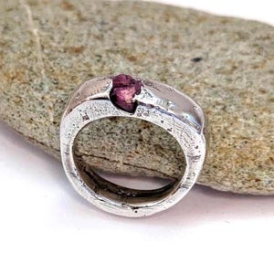 May include: A silver ring with a textured, hammered finish featuring a single, small, raw, deep pinkish-red gemstone set into the top of the band. The metal surface displays visible indentations and a rustic, uneven appearance throughout the band.