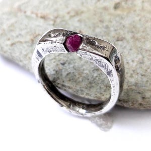 Puede incluir: Un anillo de plata con un acabado texturizado y martillado que presenta una pequeña piedra preciosa en bruto de color rojo rosado intenso engastada en la parte superior central. La superficie metálica tiene un aspecto rugoso e irregular.