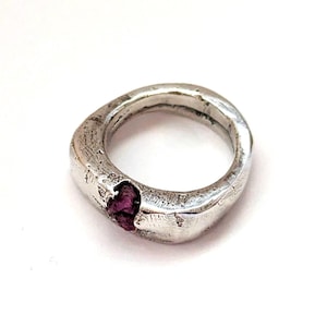 以下が含まれることがあります： ざらざらとした質感の銀色のリングで、バンドには濃いピンク色の宝石が埋め込まれています。 リングは有機的で不規則な形状をしており、宝石がアクセントになっています。
