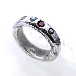 May include: A silver ring with a textured, weathered finish featuring five small, round, multi-coloured gemstones set along the outer band. The stones include shades of red, dark blue, and light purple, embedded within the uneven, oxidised metal surface.