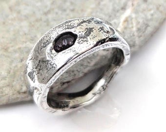 Può includere: Un anello largo in argento con una superficie fortemente testurizzata e butterata, con un inserto in pietra scura, irregolare e incassato. Il metallo presenta una finitura lucida sui bordi e un aspetto scuro e ossidato nelle cavità.