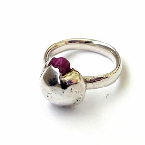 May include: A silver ring featuring a smooth, rounded, metallic setting that holds a small, raw, deep pink gemstone. The band is polished and circular, with the stone nestled within a concave, crater-like indentation on the front of the ring.