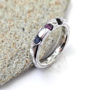 May include: A silver-toned ring with a textured, matte finish featuring three small, embedded rectangular gemstones in shades of dark blue, deep pink, and black. The band has a rugged, uneven surface texture and a smooth, polished interior surface.