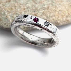 May include: A silver ring with a textured, weathered surface featuring three flush-set stones: one black stone, one vibrant red stone, and one clear stone arranged in a row along the top of the band, showcasing a rugged and organic metal finish.