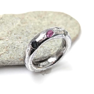 May include: A silver ring with a textured, uneven surface and a polished finish. The band features two small, embedded gemstones: one deep pink and one dark black. The interior of the band is clearly stamped with the number 925, indicating sterling silver.