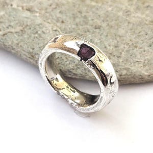 May include: A silver ring with a textured, uneven surface and a single, small, dark purple, raw-cut gemstone embedded into the top of the band. The metal has a polished finish in some areas and a rough, organic appearance throughout the band.