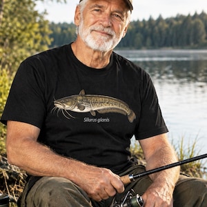 Può includere: Una t-shirt nera a maniche corte con un'illustrazione dettagliata di un siluro, con il nome scientifico Silurus glanis stampato in bianco sotto il grafico del pesce. Il capo presenta un girocollo e una vestibilità standard.