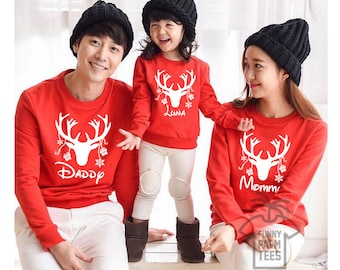 May include: A family of three wearing matching red Christmas sweaters with a white reindeer design. The sweaters have the words "Daddy", "Mommy", and "Luna" printed on them.