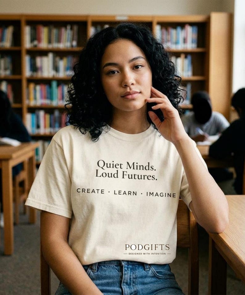 May include: A cream-colored t-shirt with the text "Quiet Minds. Loud Futures." and the words "CREATE . LEARN . IMAGINE" printed in black. The shirt also has the PODGIFTS logo at the bottom. The person is wearing blue jeans.