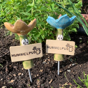 Könnte beinhalten: Zwei skurrile Gartenstecker aus Keramik mit blumenförmigen Köpfen in Beige und Blau. Jeder Stecker hat ein Schild mit der Aufschrift "HUMMELPUB" und einer Bierkrug-Grafik. Die Stecker sind in Erde gepflanzt, mit grünem Laub im Hintergrund.