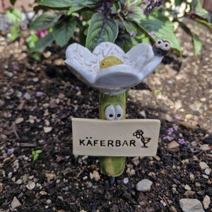 Könnte beinhalten: Ein skurriler Keramik-Gartenstecker in Form einer Blume mit einem Marienkäfer auf einem Blütenblatt. Der Stecker hat einen grünen Stiel mit Cartoon-Augen und ein Schild mit der Aufschrift "KÄFERBAR" und einer kleinen floralen Illustration.