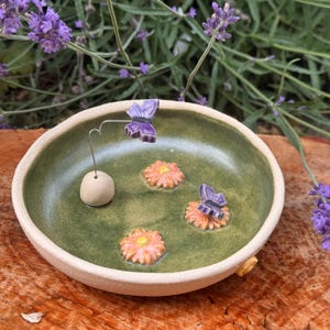 Könnte beinhalten: Eine runde Keramikschale mit moosgrünem Innenraum und hellbeiger Außenseite. Die Schale ist mit zwei kleinen geformten rosa Blumen und zwei violetten Schmetterlingsfiguren verziert, von denen eine an einem dünnen Metalldraht befestigt ist.