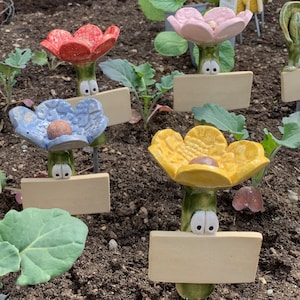 Könnte beinhalten: Fünf Keramik-Blumen-Gartenmarkierungen mit leeren Schildern. Die Blumen sind rot, blau, gelb und pink. Jede Blume hat Glitz Augen und einen Stiel.