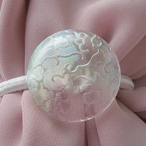 May include: A white, round, decorative curtain tieback with a textured design. The tieback is made of glass and has a white elastic band.