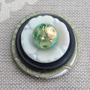 May include: A layered button with a green, iridescent center, surrounded by a white, flower-shaped layer, a black layer, and a patterned green and silver outer layer. The button is likely for sewing or crafting.