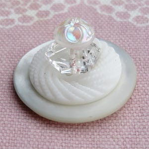 May include: A layered button with a pearlescent finish. The base is a large, flat, white button. Above it is a smaller, textured button, topped with a clear, faceted crystal. The button is set against a pink background.