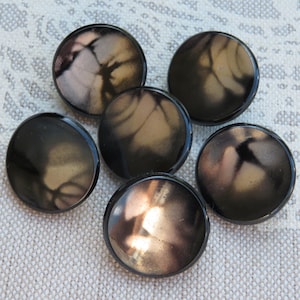 Puede incluir: Seis botones redondos de color marrón oscuro con un acabado brillante. Cada botón tiene un patrón único y arremolinado en tonos beige y negro. Los botones están dispuestos sobre una tela clara y texturizada.