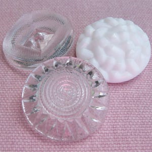 Könnte beinhalten: Drei dekorative Knöpfe auf einem rosa Stoff. Einer ist transparent mit einem strukturierten Design, ein anderer ist weiß mit einem floralen Muster und der dritte ist transparent mit einem Spiraldesign.