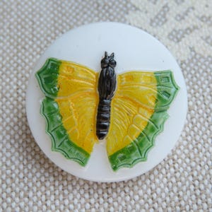 May include: A white, round decorative button with a raised butterfly design. The butterfly has yellow wings with green edges and a black body. The button is likely used for sewing or crafts.