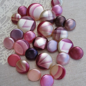 May include: A collection of round buttons in various shades of pink, mauve, and deep purple. The buttons feature a mix of solid finishes and geometric patterns, including some with white or lighter pink accents, arranged in a cluster.