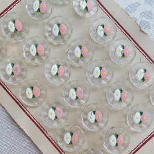 Pode incluir: Uma coleção de botões de vidro transparente facetado com desenhos florais bordados. Cada botão apresenta uma flor rosa e branca com centros amarelos. Os botões estão dispostos em um cartão de papel vintage com uma borda vermelha.