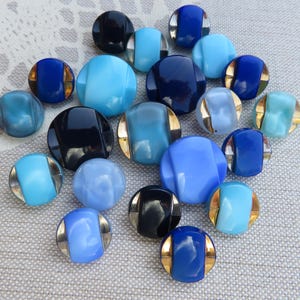 May include: A collection of round buttons in various shades of blue, light blue, and black. Many of the buttons feature a smooth, glossy finish with metallic gold-toned accents on the sides, creating a distinct contrast against the blue and black centres.